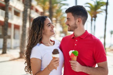 Young hispanic couple tourists hugging each other eating ice cream at seaside