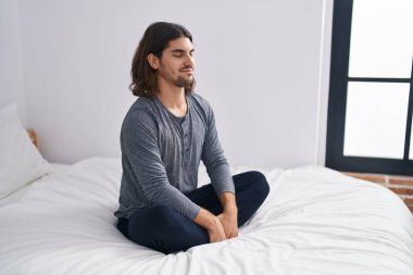 Young hispanic man sitting on bed meditating at bedroom