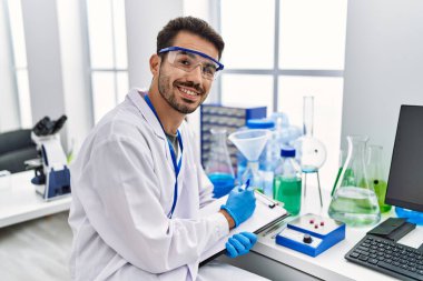 Young hispanic man wearing scientist uniform writing on clipboard working at laboratory