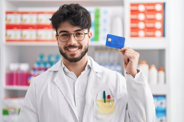 西班牙裔男子 留着胡子 在药店工作 手里拿着信用卡 站在那里 面带微笑 脸上带着自信的笑容 露出牙齿 — 图库照片