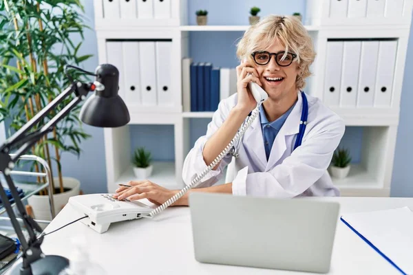 Jovem Loiro Homem Médico Usando Laptop Falando Telefone Clínica — Fotografia de Stock