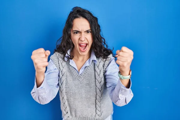 青の背景に立っている若いブルネットの女性は怒りで叫んでいる間に怒りと怒りに苛まれ 激怒した拳を上げる 怒りと攻撃的な考え方 — ストック写真