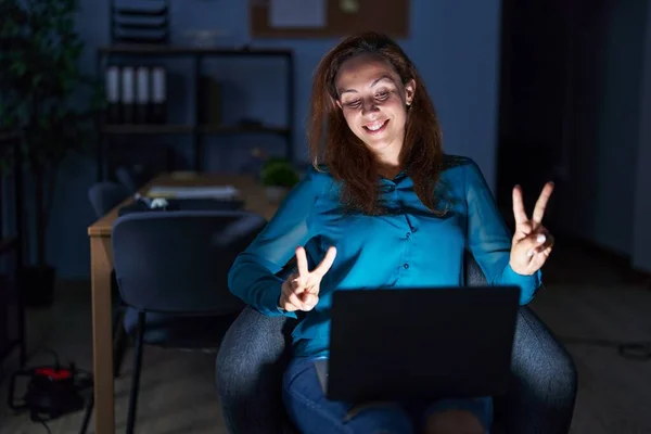 Brunette woman working at the office at night smiling looking to the camera showing fingers doing victory sign. number two.