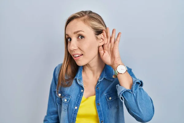 若いブロンドの女性が青い背景の上に立ち 噂やゴシップに耳を傾けながら手で笑っている 聴覚障害の概念 — ストック写真