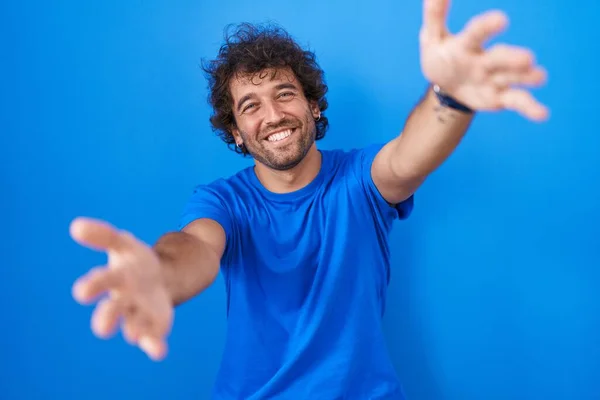 カメラを見て青の背景に立ってヒスパニック系の若い男が抱擁のために腕を開いて笑顔 幸せを受け入れる陽気な表情 — ストック写真