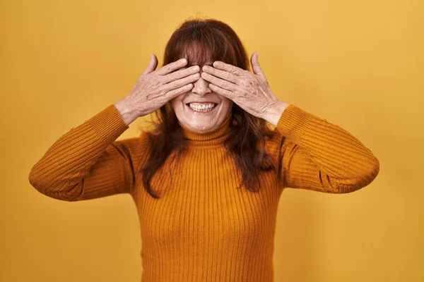 黄色の背景に手を明るく面白い笑顔で目をカバー上に立っている中年のパニック女性 視覚障害の概念 — ストック写真