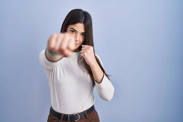 Young brunette woman standing over blue background punching fist to fight, aggressive and angry attack, threat and violence