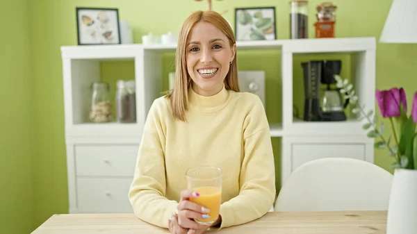 年轻的金发女人坐在家里的桌上喝橙汁 — 图库照片