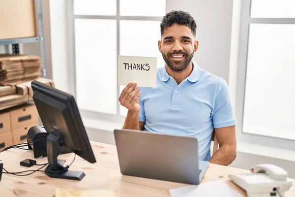 Hispanic man with beard working at the office with laptop holding thanks banner looking positive and happy standing and smiling with a confident smile showing teeth