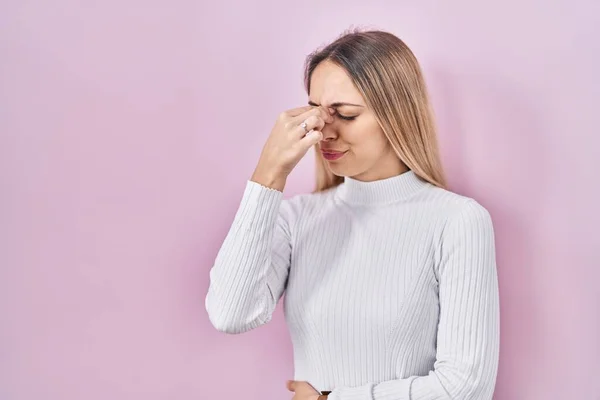若いブロンドの女性はピンクの背景に白いセーターを着て疲れ鼻をこすり 目は疲労と頭痛を感じます ストレスとフラストレーションの概念 — ストック写真