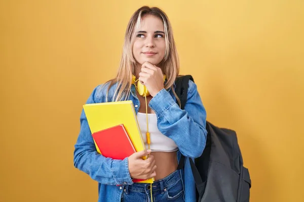 若いブロンドの女性学生のバックパックを着て 質問について考える顎に手で本を保持し 集中的な表現 笑顔で思いやりのある顔 疑わしい概念 — ストック写真