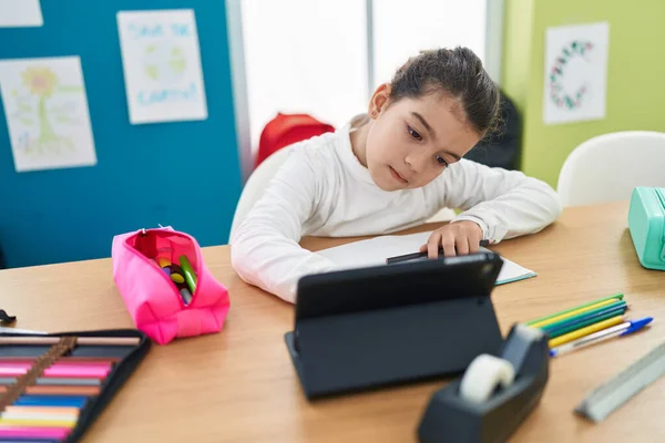 Adorable hispanic girl student writing on notebook using touchpad at classroom