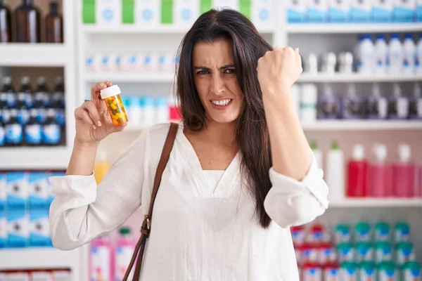 Young brunette woman shopping at pharmacy drugstore holding pills annoyed and frustrated shouting with anger, yelling crazy with anger and hand raised