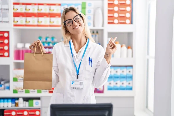 Joven Mujer Rubia Trabajando Farmacia Sosteniendo Bolsa Papel Sonriendo Feliz —  Fotos de Stock