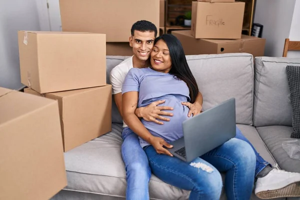 Young latin couple expecting baby touching belly using laptop at new home