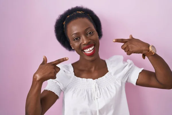 头发卷曲的非洲妇女站在粉色的背景上 笑容满面 用手指 牙齿和嘴指指点着 牙齿健康概念 — 图库照片