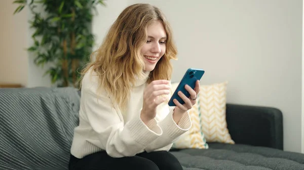 年轻的金发女人坐在家里的沙发上用智能手机 — 图库照片