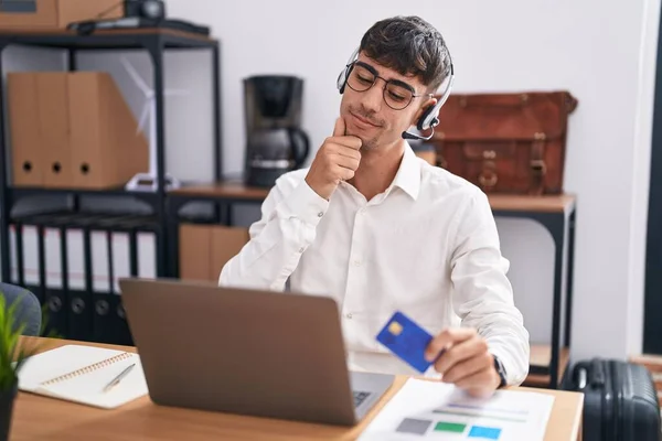 stock image Young hispanic man working using computer laptop holding credit card with hand on chin thinking about question, pensive expression. smiling and thoughtful face. doubt concept. 