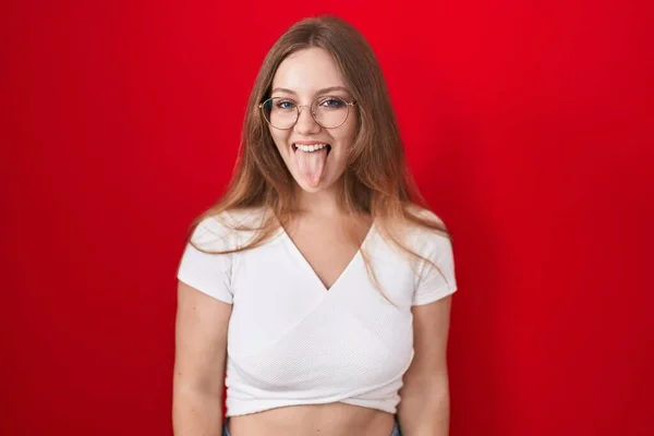 年轻的高加索女人站在红色的背景上 带着滑稽的表情伸出舌头 高兴极了 情感概念 — 图库照片