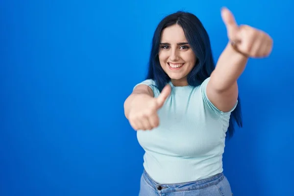 Young modern girl with blue hair standing over blue background approving doing positive gesture with hand, thumbs up smiling and happy for success. winner gesture.