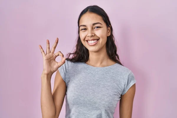 年轻的巴西女人穿着宽松的T恤 粉红的背景 微笑着积极地做着 手指手画脚地签了名 成功表达 — 图库照片