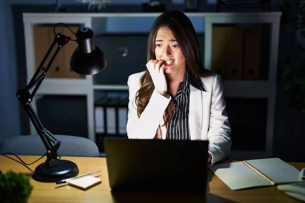 若いブルネットの女性は ノートパソコンで夜にオフィスで働いており 口の爪をかむ上で手でストレスと緊張しています 不安の問題 — ストック写真