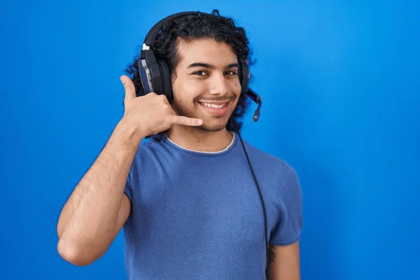 头发卷曲的西班牙裔男人用耳机听音乐 微笑着用手和手指做电话手势 就像在电话里说话一样 交流概念 — 图库照片