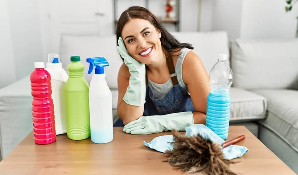 年轻美丽的惊慌失措的女人面带微笑 自信地靠着桌子 在家里放着干净的产品 — 图库照片