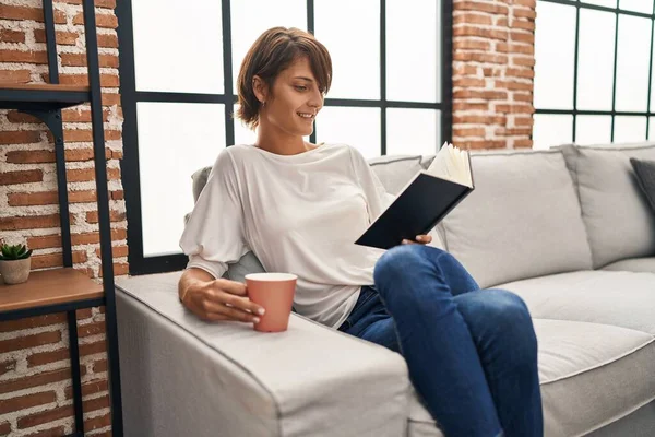 Young Beautiful Hispanic Woman Reading Book Drinking Coffee Sitting Sofa — Stock fotografie