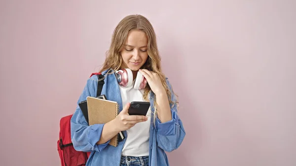 若いですブロンド女性学生ホールディング本を使用してスマートフォンで疑い式上の隔離されたピンクの背景 — ストック写真