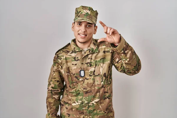 迷彩軍の制服を着た若いアラブ人男性が笑顔で 指を見てカメラで小さなサイズのサインをする手で自信を持ってジェスチャー 概念を測定し — ストック写真
