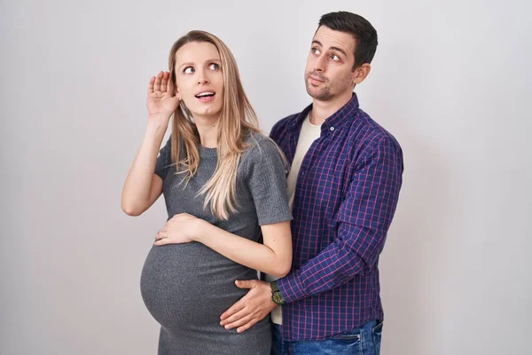 噂やゴシップに耳を傾ける耳の上に手で笑みを浮かべて白い背景に立って赤ちゃんを期待若いカップル 聴覚障害の概念 — ストック写真