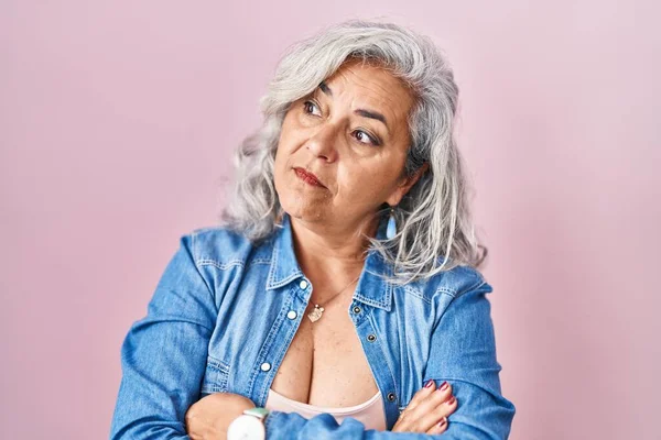 Middle age woman with grey hair standing over pink background looking to the side with arms crossed convinced and confident