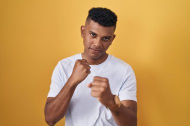 Young hispanic man standing over yellow background ready to fight with fist defense gesture, angry and upset face, afraid of problem 