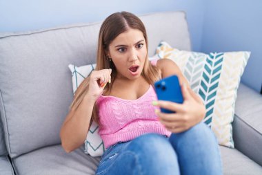 Kanepede oturan akıllı telefonlu genç sarışın kadın korkmuş ve ağzı açık bir şekilde şaşkınlık içinde bekliyor. 
