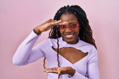 Pembe arka planda örülü saçları olan Afrikalı bir kadın. Elleri büyük ve büyük boylarda işaret ediyor. Ölçü sembolü. Kameraya bakıp gülümsüyor. ölçüm kavramı. 