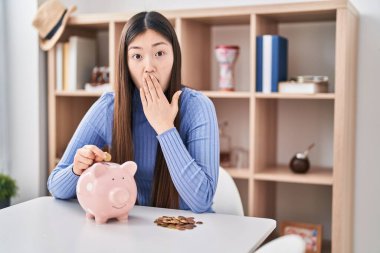 Çinli genç bir kadın domuz kumbarasına para koyuyor ağzını eliyle kapatıyor, şok geçiriyor ve hata yapmaktan korkuyor. şaşırmış ifade 