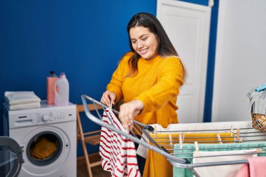 Young beautiful plus size woman smiling confident hanging clothes on clothesline at laundry room