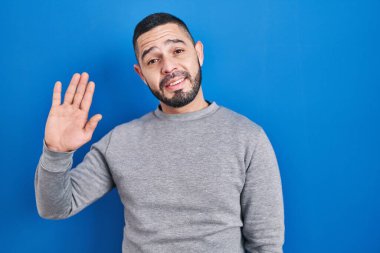 Hispanic man standing over blue background waiving saying hello happy and smiling, friendly welcome gesture 