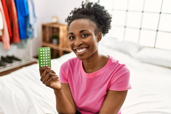 African american woman holding birth control pills sitting on bed at bedroom