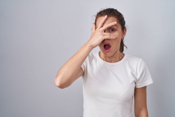 Красивая брюнетка женщина, стоящая на изолированном фоне, подглядывая в шокирующие закрывающие лицо и глаза рукой, глядя сквозь пальцы со смущенным выражением лица. 