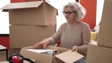 Middle age woman with grey hair unpacking cardboard box reading document at new home