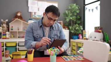 Young chinese man preschool teacher using touchpad sitting on table at kindergarten