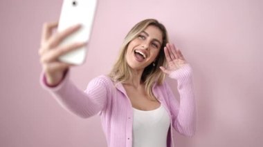 Genç sarışın kadın kendinden emin gülümsüyor. Pembe arka planda akıllı telefondan selfie çekiyor.