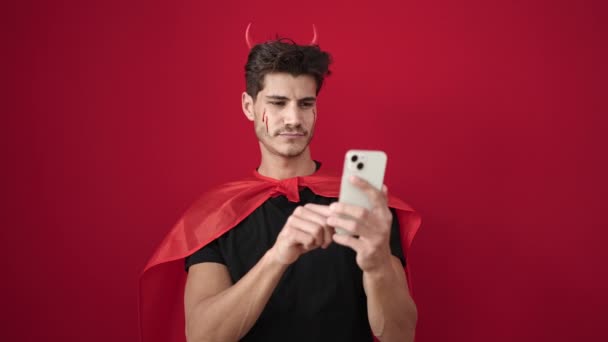 若いですヒスパニック男身に着けています悪魔衣装使用スマートフォン上の隔離された赤の背景 — ストック動画