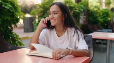 Genç, İspanyol asıllı, kitap okuyan, kahve dükkanının terasında akıllı telefondan konuşan bir kadın.