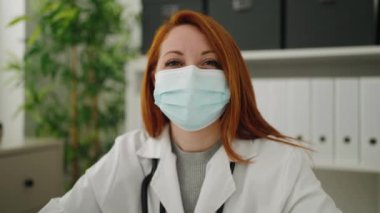 Young redhead woman wearing doctor uniform and medical mask working at clinic