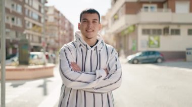 Genç adam kendinden emin gülümsüyor. Kollarını kavuşturup caddede hareketler yapıyor.