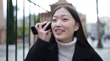 Young chinese woman smiling confident listening audio message by the smartphone at street