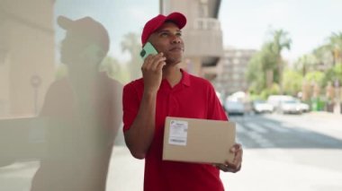 Genç latin teslimat işçisi elinde paketle caddede akıllı telefondan konuşuyor.